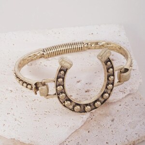 May include: A gold-toned metal bracelet featuring a central horseshoe charm adorned with small raised circular studs. The band consists of a textured metal segment and a coiled spring section, connected to the horseshoe by hinged links.