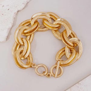 May include: A chunky gold-toned chain link bracelet featuring large, textured oval links with a matte finish. The bracelet is secured with a circular toggle clasp and a matching T-bar closure, creating a bold and substantial piece of jewelry.