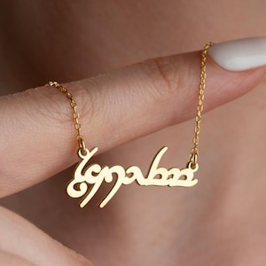 May include: A gold-colored necklace featuring a delicate chain and a central pendant displaying custom text written in a stylized, flowing script. The metal has a polished finish, and the pendant is suspended from a thin, matching gold-tone link chain.