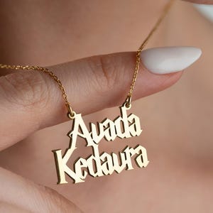 May include: A gold-toned metal necklace featuring a pendant with the text Avada Kedaura written in a stylized gothic font. The pendant is suspended from a delicate gold-toned chain that rests against the neck, showcasing a polished finish.