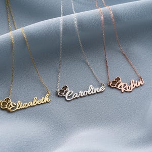 May include: Three personalized name necklaces in gold, silver, and rose gold. Each necklace features a name pendant with a crown design. The names are "Elizabeth", "Caroline", and "Robin". The necklaces are displayed on a light blue fabric.