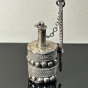 May include: A cylindrical silver-toned metal container with a narrow neck and a stopper attached by a small chain. The body features horizontal bands of twisted rope-like texture alternating with rows of small, raised spherical metal beads.