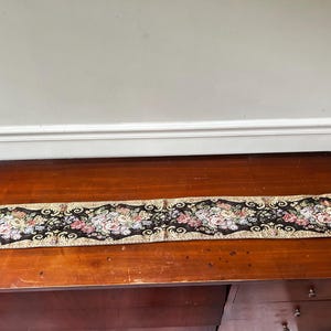 May include: A long, rectangular tapestry wall hanging featuring a black background with a repeating floral pattern in shades of pink, white, and green. The tapestry is framed by gold-colored metal end caps with circular rings on each side.