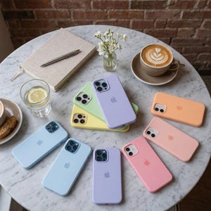 May include: A collection of silicone smartphone cases in various pastel colors including lavender, light blue, yellow, peach, and pink. Each case features a camera cutout and the Apple logo on the back. The cases are arranged on a round marble surface.