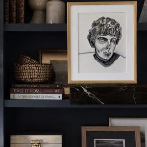 May include: A framed black and white ink portrait of a person with curly hair, displayed in a gold frame with a white mount. The artwork features expressive facial features and is positioned on a dark shelf alongside books and decorative objects.