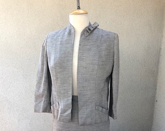 May include: A gray skirt suit with a ruffled collar on a mannequin. The jacket has a mandarin collar and three-quarter sleeves. The skirt is knee-length.