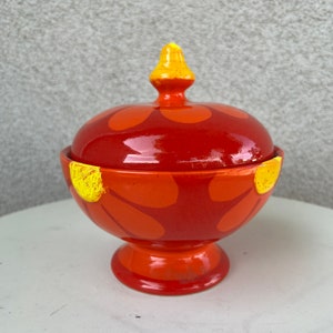Könnte beinhalten: Eine kleine, orangefarbene Zuckerdose aus Keramik mit Deckel. Die Schale hat ein gelbes und orangefarbenes Blumenmuster. Der Deckel hat einen kleinen, gelben Knopf.