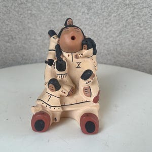 May include: A hand-painted ceramic figurine of a Native American woman with a large family. The woman is wearing a traditional dress and has a large, round face. She is holding a small child in her arms, and there are other children around her. The figurine is painted in shades of brown, black, and white.