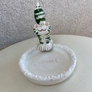 Può includere: Figurina di gatto in ceramica bianca con strisce verdi, seduta con la testa inclinata all'indietro. Il gatto tiene un piatto in ceramica bianca con un disegno a lisca di pesce.