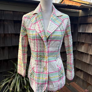 May include: A vintage blazer in a plaid pattern of pink, green, yellow, and white. The jacket has a notched collar, two front pockets, and three yellow buttons. The blazer is displayed on a mannequin.