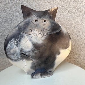 Op de afbeelding: Een keramische kattenbeeldhouwwerk met een grillig ontwerp. De kat is voornamelijk grijs en wit, met zwarte accenten en een lachend gezicht. Het beeldhouwwerk heeft een afgerond lichaam en puntige oren en is een uniek kunstwerk.