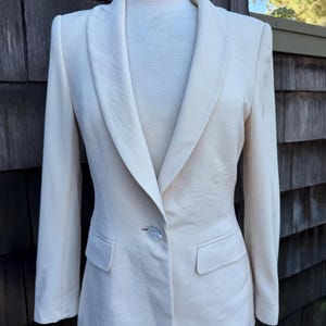 May include: A cream-colored blazer featuring a shawl collar, long sleeves, and a single-button closure at the front. The garment includes two horizontal flap pockets positioned at the waistline and is displayed on a mannequin torso.