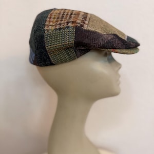 May include: A patchwork newsboy cap in shades of brown, green, and blue. The cap features a variety of textured fabrics, including tweed and plaid, sewn together in a unique pattern. The brim is a solid brown color.