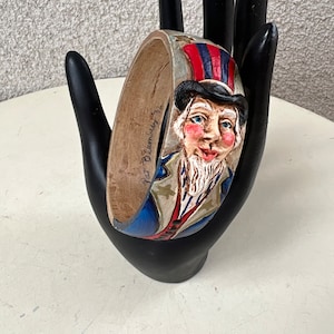 May include: A wooden bangle bracelet featuring a carved Uncle Sam figure. The bracelet is painted with red, white, and blue accents, and the Uncle Sam figure has a top hat. The bracelet is held by a black hand.