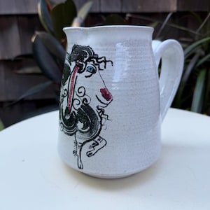 May include: White ceramic pitcher with a whimsical illustration of a creature with black, red, and green details. The pitcher has a handle and a slightly textured surface, perfect for serving beverages.
