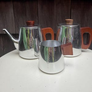 May include: A set of three vintage silver-toned tea or coffee serving pieces. The set includes two teapots and a creamer, each with wooden handles and lids. The teapots have a decorative leaf design. The pieces are arranged on a white surface.