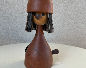 以下が含まれることがあります： 茶色の帽子をかぶり、黒い髪と茶色の体をした木製の置物。置物は丸い顔に黒い目と茶色の鼻をしています。置物は右手につまようじを持っています。