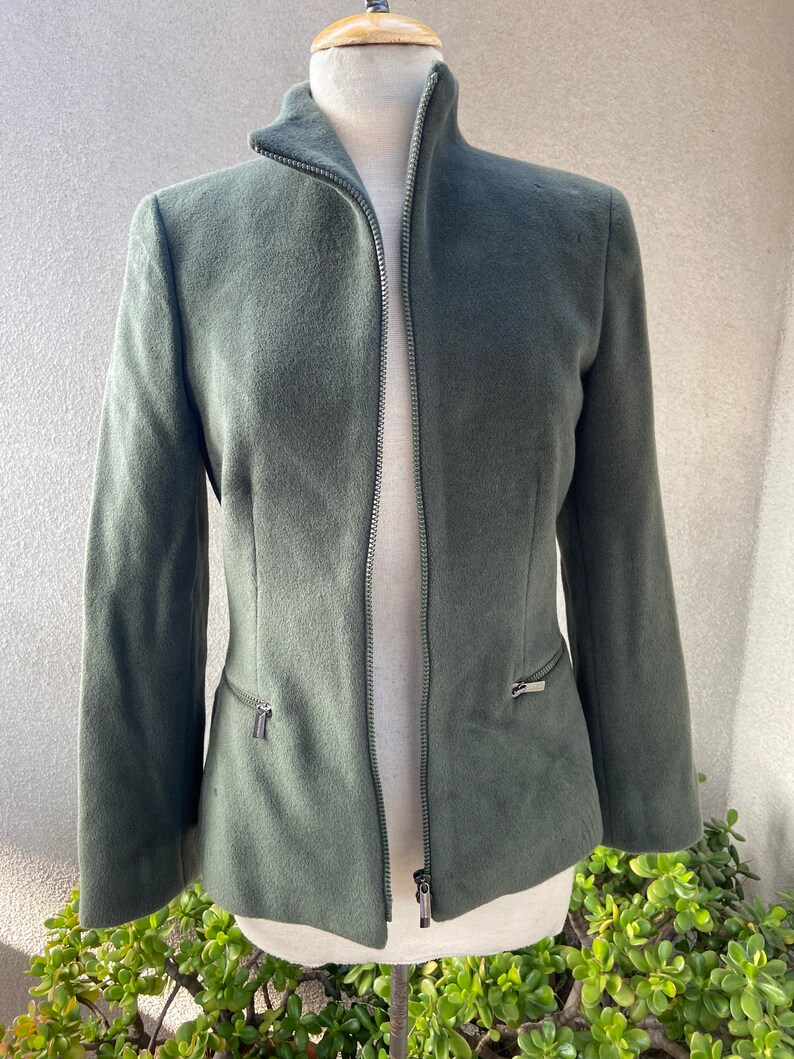 May include: A dark green, zippered jacket with a high collar. The jacket has two zippered pockets on the sides.