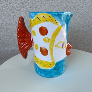 May include: A blue ceramic pitcher with a fish design. The fish is painted in yellow, orange, and white with black eyes and a red mouth. The pitcher has a handle shaped like a fish tail.