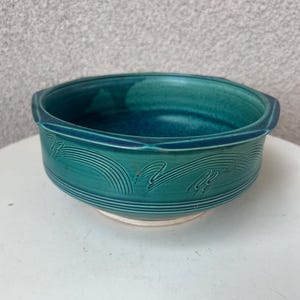 May include: A teal ceramic bowl with a wide rim and a white base. The bowl has a glossy finish and is decorated with curved lines. The bowl is approximately 8 inches in diameter and 3 inches tall.