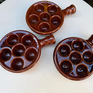 May include: Three brown ceramic escargot dishes with handles. Each dish has seven circular indentations for holding snails. The dishes are glossy and arranged on a white surface.