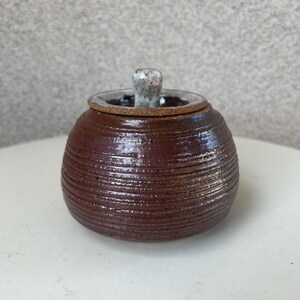 May include: A small, round, brown ceramic jar with a white knob lid. The jar has a textured, ribbed surface.