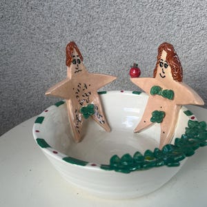 May include: A white ceramic bowl with green and red trim. Two ceramic figures of people are in the bowl. The figures are nude and have green leaves around their waists. One figure is holding a red apple.