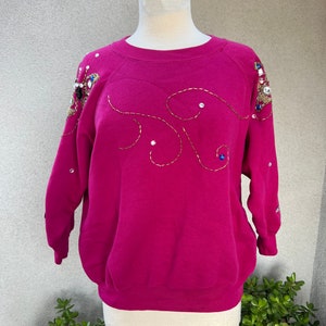 Puede incluir: Una sudadera magenta con un diseño decorativo de hilo dorado y pedrería en la parte delantera y las mangas.