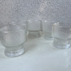 Könnte beinhalten: Fünf klare Glas-Trinkgläser mit einer strukturierten Oberfläche. Die Gläser sind alle gleich groß und haben die gleiche Form und sind in einer Reihe auf einer weißen Oberfläche angeordnet.