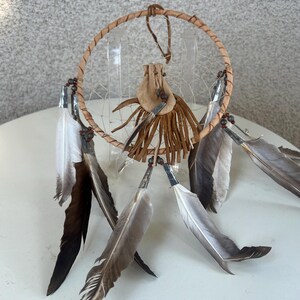 May include: A brown leather dreamcatcher with a small pouch and feathers hanging from the bottom. The dreamcatcher is made with a wooden hoop and a web of string.