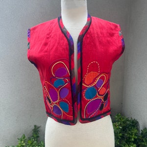 May include: A red sleeveless vest with a black trim and colorful embroidered flowers. The vest has a V-neck and a front closure.