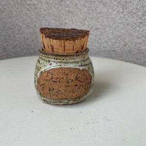 May include: A small, brown and white ceramic jar with a cork lid. The jar has the word "remarkable" written on it.