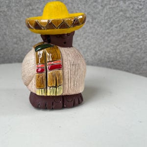 Puede incluir: Figura de cerámica de una persona con sombrero amarillo y bufanda colorida. La figura tiene una cara marrón y viste una camisa color crema. La base de la figura es de color marrón oscuro.