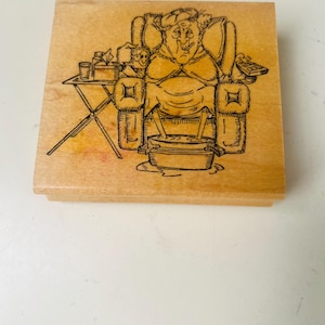 Peut inclure: Un tampon en bois avec une illustration de dessin animé d'une personne se relaxant dans un fauteuil avec un bain de pieds. La personne tient une télécommande. Une petite table avec une boisson et des mouchoirs est à côté de la chaise. Le tampon est sur un bloc de bois clair.