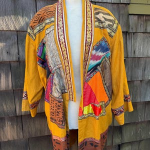 May include: A mustard yellow open-front jacket featuring a patchwork design with various patterns including animal prints, geometric shapes, and floral motifs. The garment has long sleeves and a decorative border along the collar and front opening.