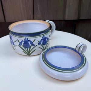 Puede incluir: Un cuenco de cerámica para sopa con tapa a juego, que presenta un patrón floral de lirios azules pintado a mano sobre un fondo blanco. El cuenco tiene un asa lateral y la tapa incluye un pequeño asa de lazo, ambos con bandas azules y verdes.