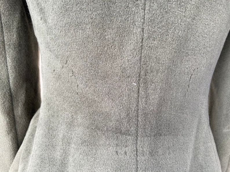 May include: Close-up of the back of a gray wool coat. The coat has a seam down the center back and two seams on either side of the center seam. The seams are visible because they are stitched with a contrasting thread.