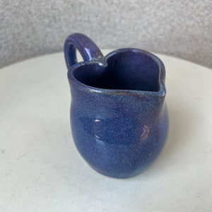 May include: A small, heart-shaped ceramic creamer in a deep blue color with a speckled glaze. The creamer has a handle and a rounded base.