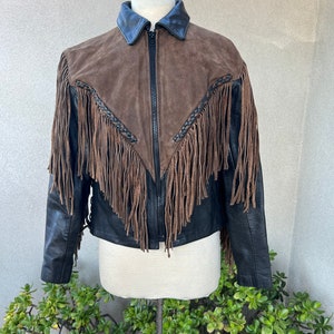 Puede incluir: Una chaqueta de cuero marrón y negro con detalles de flecos. La chaqueta tiene una cremallera y un cuello.
