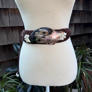 May include: A brown macrame belt with a large, iridescent shell centerpiece. The shell is surrounded by small white floral accents. The belt is displayed on a mannequin, showcasing its design and texture. The belt is a unique fashion accessory.