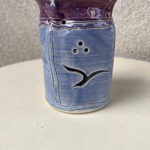 Puede incluir: Un pequeño portavelas de cerámica azul y morado con un diseño de pájaro recortado. El portavelas tiene una superficie texturizada y varios agujeros pequeños.