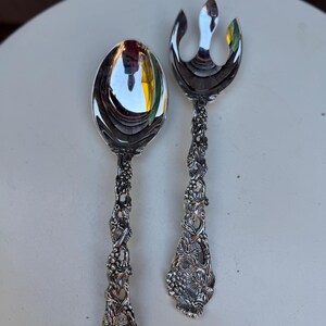 Könnte beinhalten: Ein silbernes Servierlöffel- und Gabel-Set. Der Löffel hat eine große, ovale Schüssel, und die Gabel hat drei Zinken. Beide Utensilien verfügen über verzierte Griffe mit einem Trauben- und Weinranken-Design. Das Set ist wahrscheinlich zum Servieren von Salat oder anderen Gerichten.