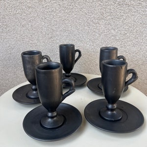 May include: Set of six black ceramic espresso cups and saucers. The cups have a classic design with a handle and a flared base. The saucers are round and have a slightly raised edge.