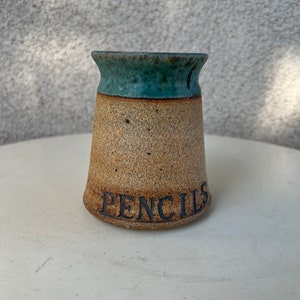 May include: A small, brown and green ceramic pencil holder with the word "PENCILS" inscribed on the front.