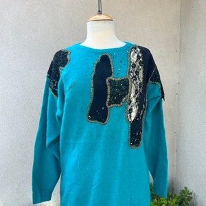 May include: A turquoise blue sweater with black and gold embellishments. The sweater has a round neckline and long sleeves.