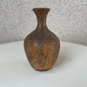 Könnte beinhalten: Eine kleine, braune Keramikvase mit einer strukturierten Oberfläche. Die Vase hat einen schmalen Hals und einen runden Körper.
