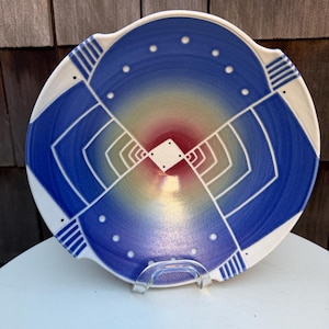 May include: A decorative ceramic plate with a blue and white geometric design. The plate features a gradient of colors, including blue, red, and yellow. The plate is supported by a clear plastic stand.