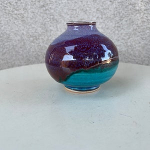 Può includere: Un piccolo vaso rotondo in ceramica con una smaltatura viola, blu e verde. Il vaso ha un'ampia apertura e una base stretta.