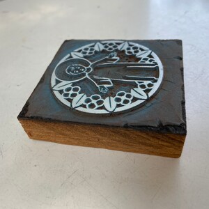 May include: A wooden block with a blue and white design of a person in a circle surrounded by leaves. The design is raised and appears to be a stamp.