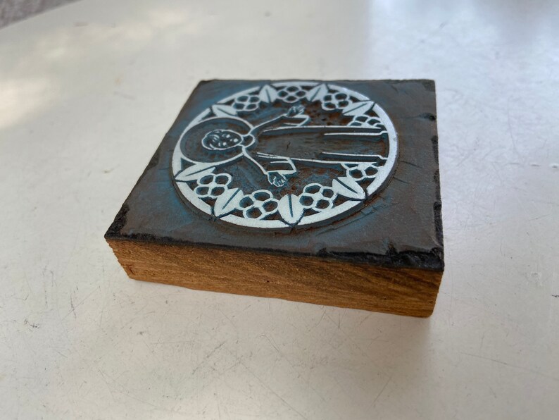 May include: A wooden block with a blue and white design of a person in a circle surrounded by leaves. The design is raised and appears to be a stamp.